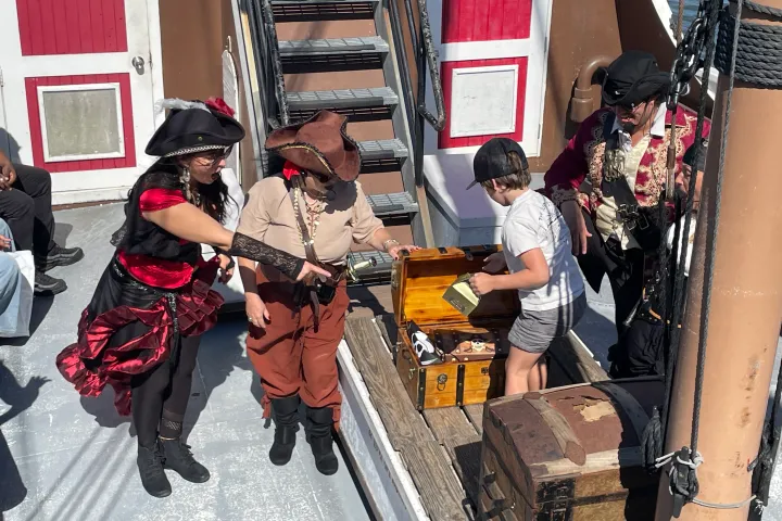 Pirate-themed scene with adults and child opening wooden treasure chest on a ship deck.