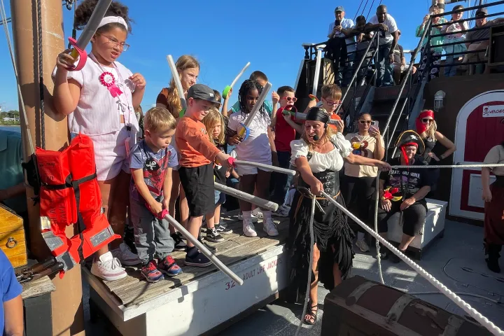 Group of children and adults dressed as pirates on a boat, holding toy swords and cheering.
