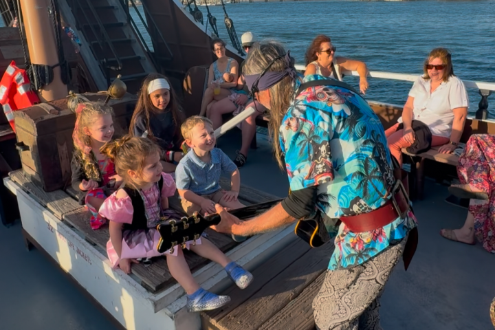 Musician playing guitar for children dressed as pirates on a boat with adults watching.