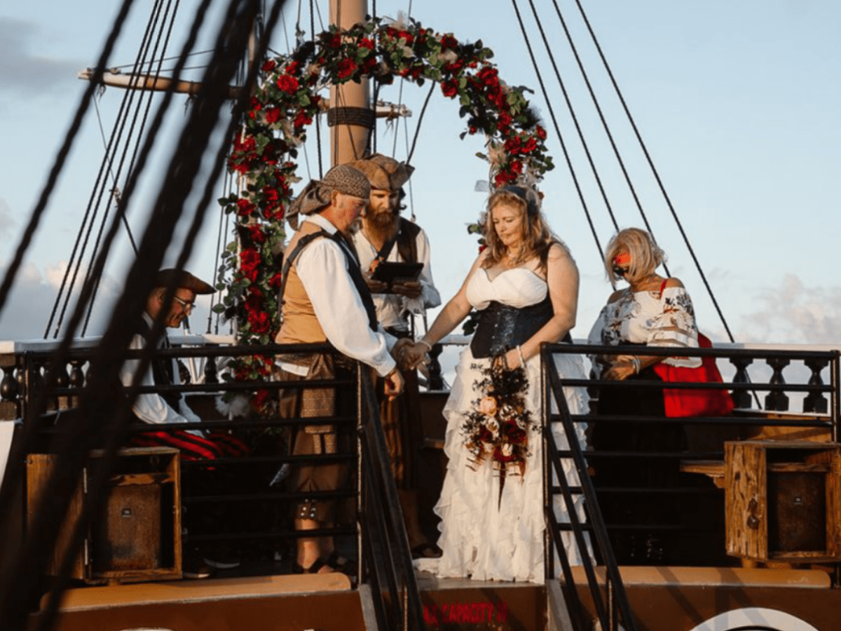 Pirate-themed wedding on a ship with bride and groom in costume under a flower arch.
