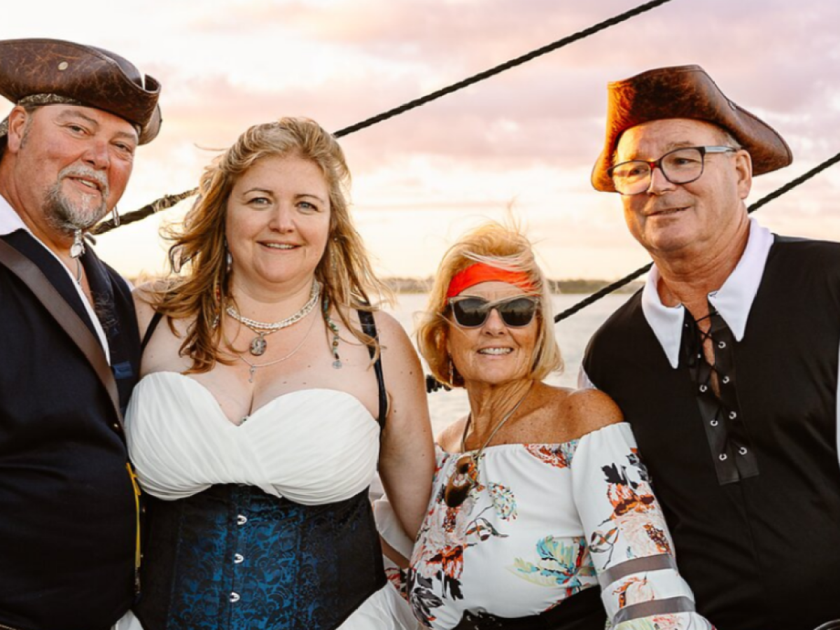 Four people in pirate costumes smiling on a boat during sunset.