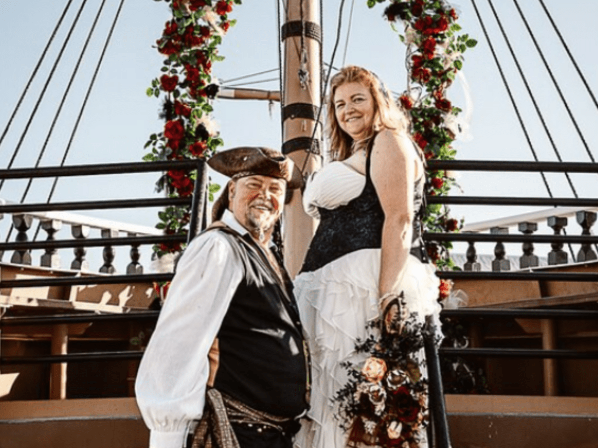 Couple in pirate-themed attire on a ship under a floral arch with a mast in the background.