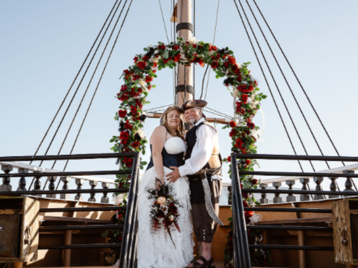 Couple in wedding attire on ship deck under an arch decorated with red and white flowers.
