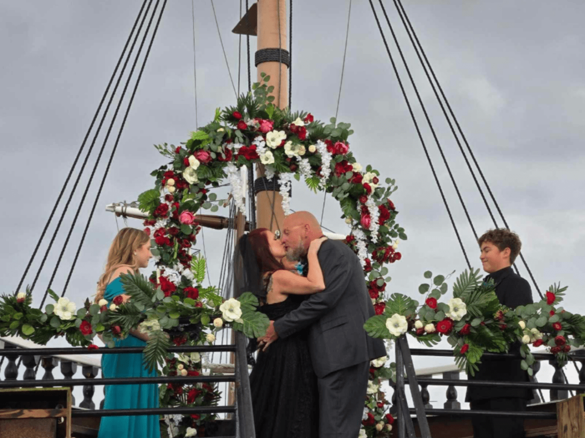 Couple kissing on a decorated ship with floral arch and two people standing by.