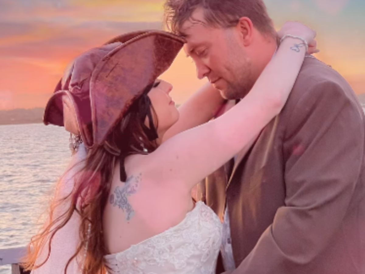 Couple embracing by the water at sunset, woman wearing a tricorn hat.
