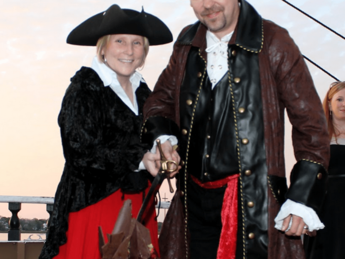 Two people in pirate costumes cutting a chocolate cake with a sword on a ship deck.