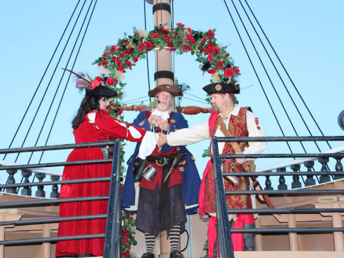 Three people in pirate costumes on a ship, with floral arch, shaking hands.