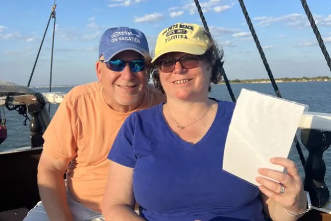 Couple on a boat, seated, both wearing hats, holding papers. Sunny day with blue sky and few clouds.