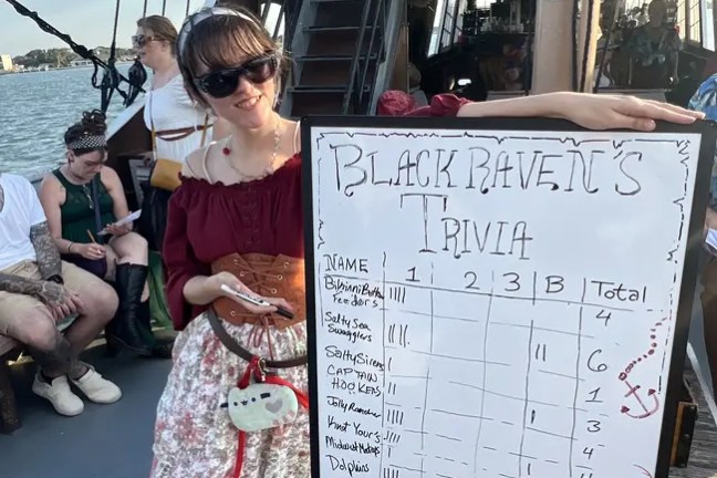 Woman on a boat holds a trivia scoreboard with people in the background.