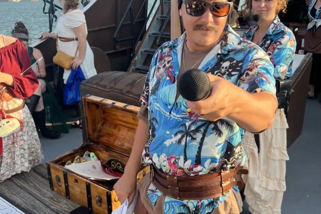 Person in pirate outfit with floral shirt on ship holding a microphone towards the camera.