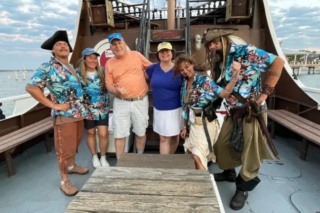 Group on a boat dressed in pirate-themed attire, smiling against a cloudy sky background.