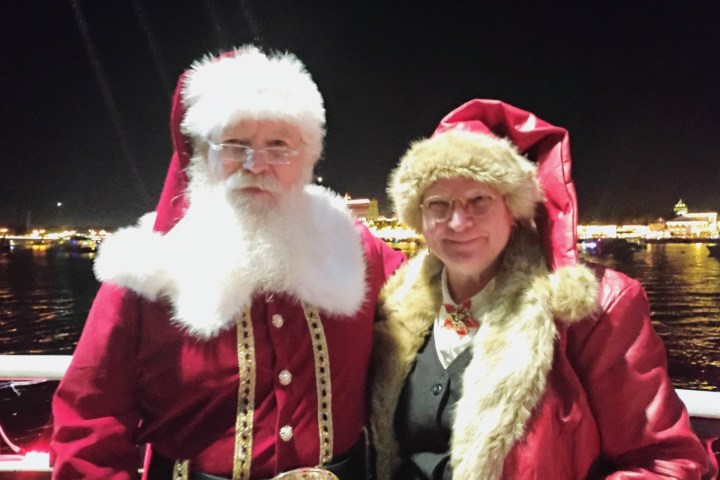 Two people in Santa outfits at night on a boat with city lights in the background.