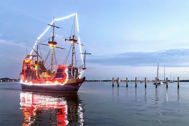 Illuminated tall ship at sea near a harbor with a blue sky.