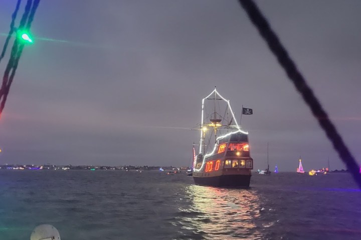 Ship with colorful lights sails on water at dusk, other lit boats in background.